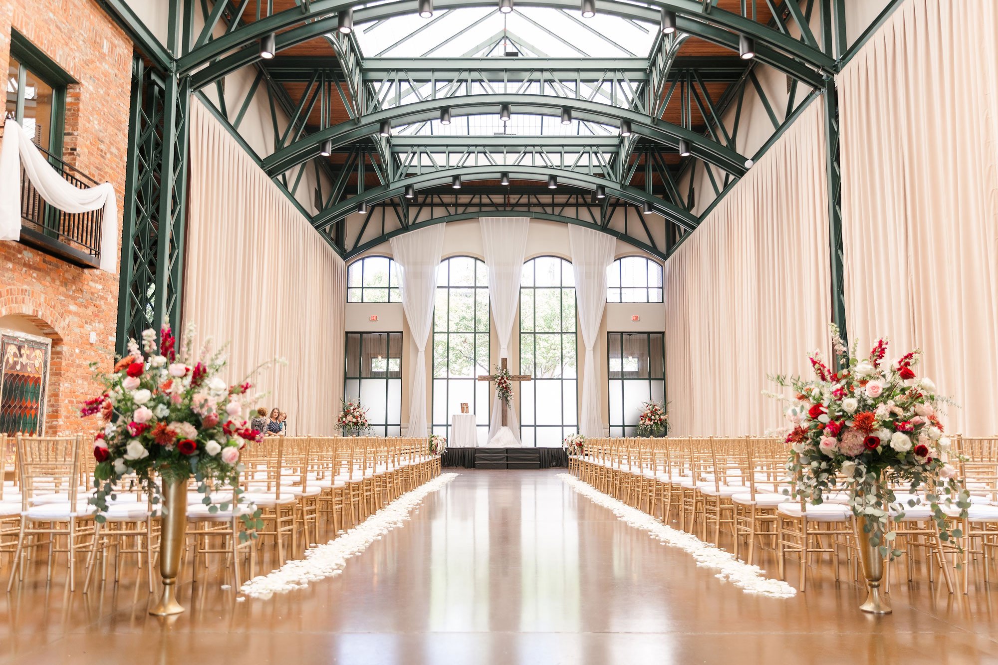 A wedding ceremony setup in an Anderson, SC event venue, featuring gold chairs, white floral arrangements on pedestals, and a cross centered in front of a tall window.