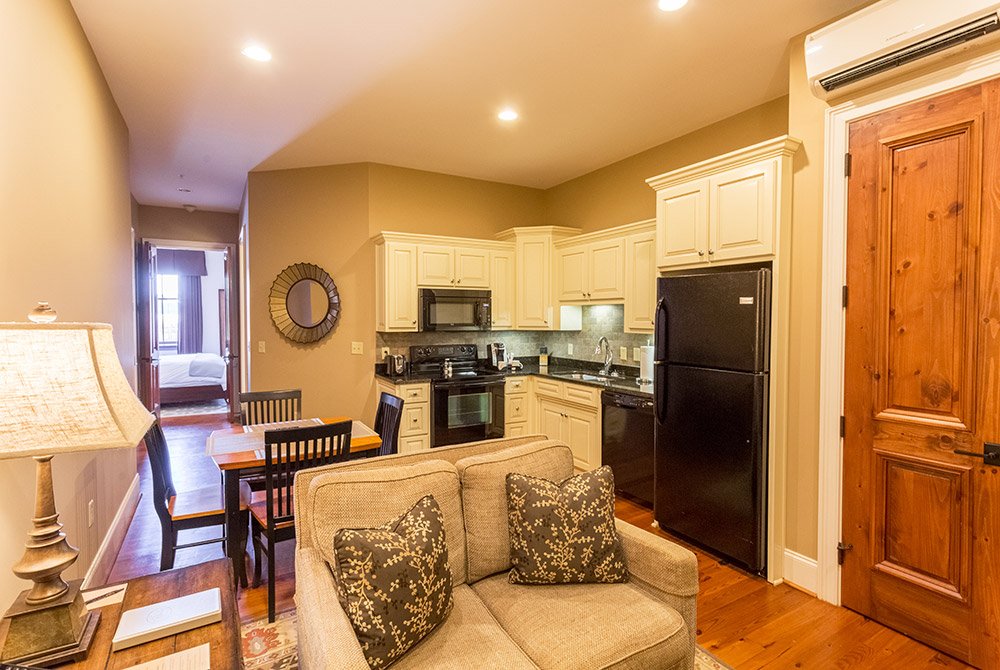 A kitchen with light-colored cabinets, black appliances, and a dining table, viewed from an adjacent living area with a sofa in one of our extended stay suites in Anderson SC.