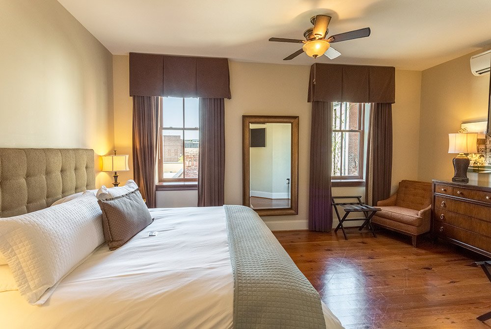 A hotel room with a king bed, a large mirror, an armchair, and a wooden dresser on a hardwood floor, typical of extended stay suites in Anderson SC.