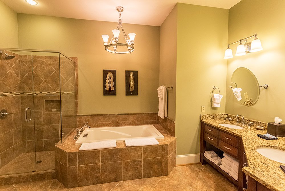 Bathroom with a glass-enclosed shower, a tiled bathtub with two towels, and a double vanity with a granite countertop and oval mirror in our extended stay suites in Anderson SC.