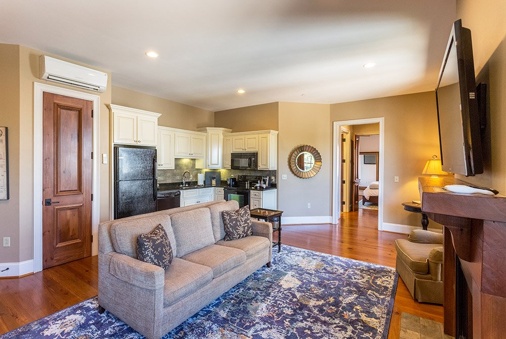A living room with a beige sofa facing a television above a fireplace, open to a kitchen with white cabinets and black appliances in one of our extended stay suites in Anderson SC.