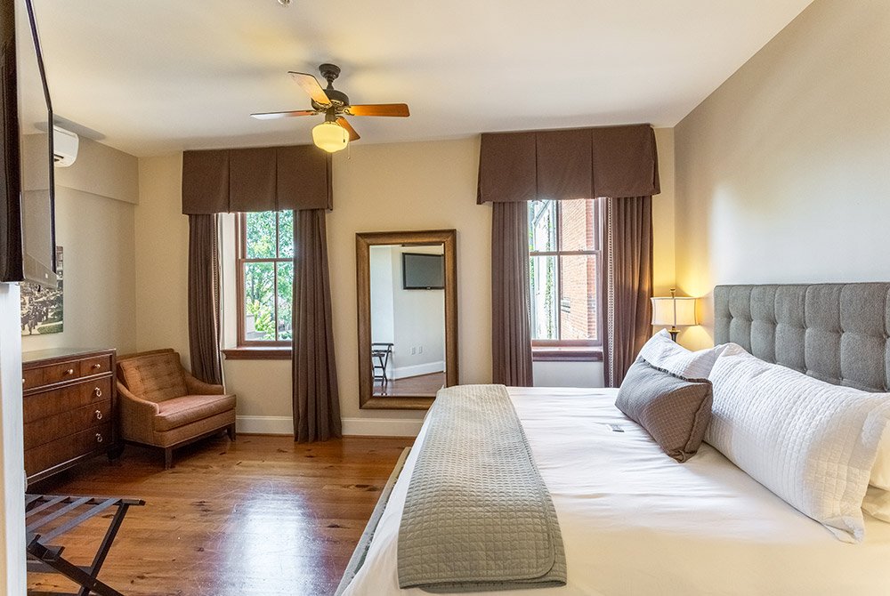 A hotel room in one of our extended stay suites in Anderson SC, featuring a queen bed, floor mirror, armchair, and dresser on wood floors.