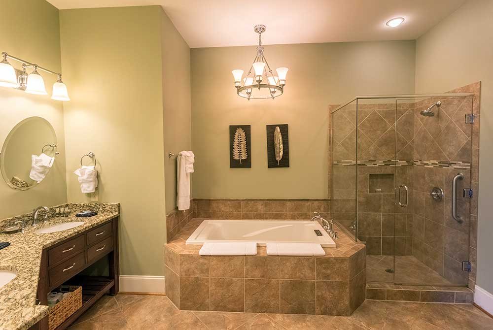 A bathroom with sage green walls, a granite-topped vanity, a glass-enclosed shower, and a jetted tub set in a tile surround, typical of extended stay suites in Anderson, SC.