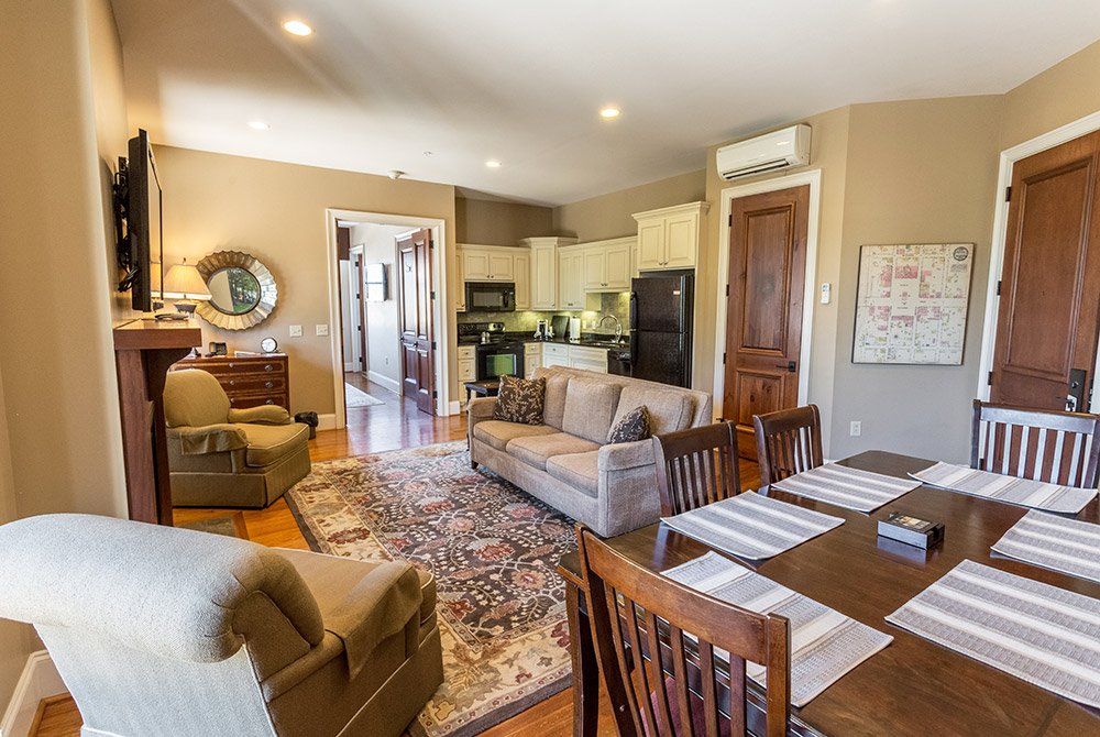An open-concept living area in our extended stay suites in Anderson, SC, featuring a sofa, two armchairs, a dining table for four, and a view into a kitchen with light-colored cabinets.