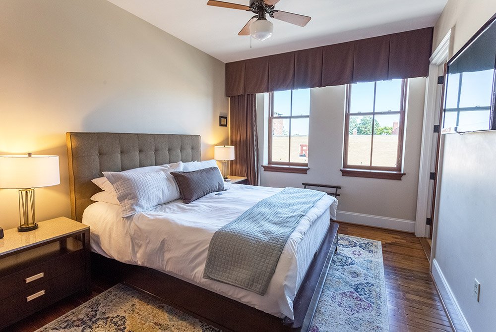 A hotel room among extended stay suites in Anderson SC, featuring a queen-sized bed with white bedding, two bedside lamps, a ceiling fan, and windows with brown curtains.