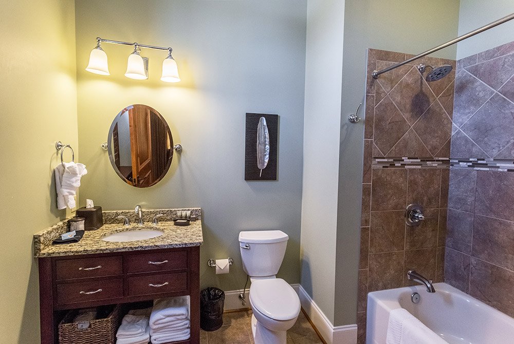 Bathroom in our extended stay suites in Anderson, SC, featuring a dark wood vanity with a stone countertop, an oval mirror, a toilet, and a tiled shower-tub combination.