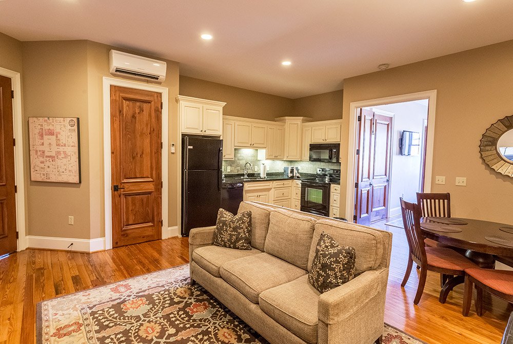 A living room with a beige sofa, patterned rug, and hardwood floors opens into a kitchen with white cabinets, black appliances, and a dining area, typical of extended stay suites in Anderson, SC.
