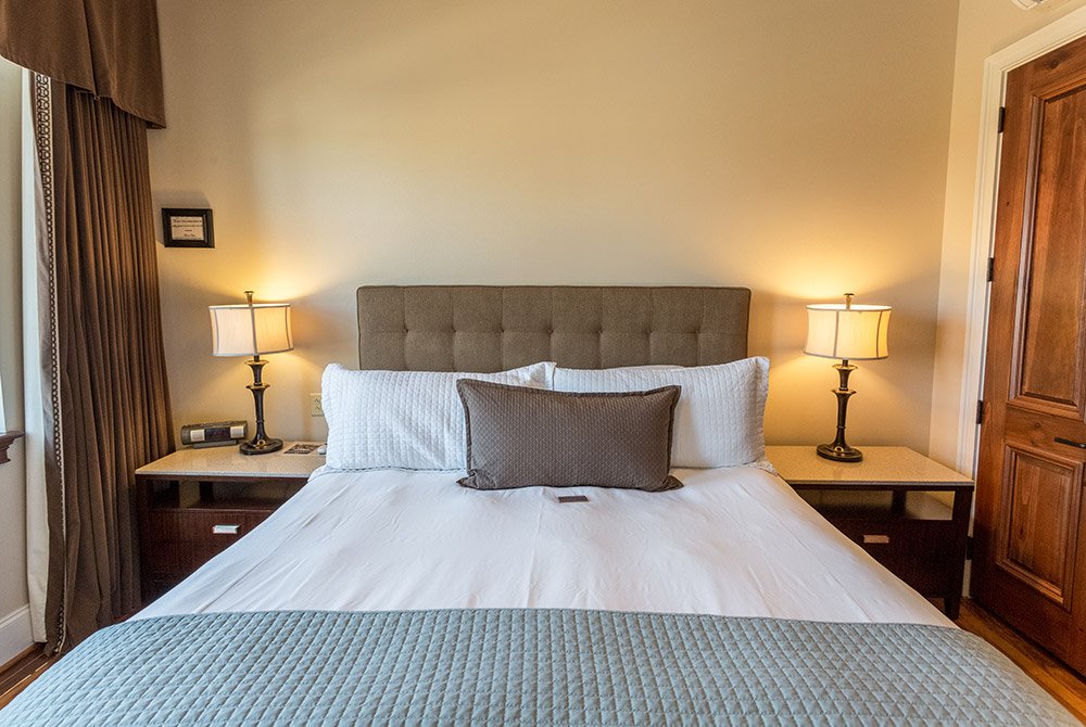 A hotel bed with white linens and a gray runner, flanked by two wooden nightstands with lit lamps in these extended stay suites in Anderson SC.