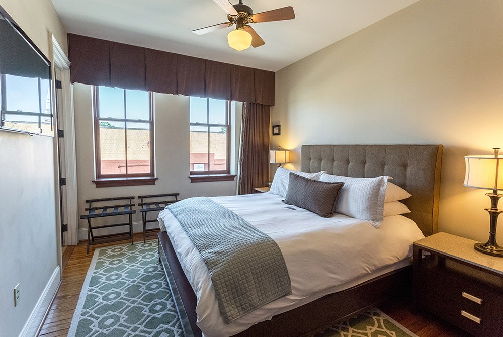 A hotel room at these extended stay suites in Anderson, SC, featuring a queen bed, two windows with brown valances, and a television mounted on the wall.
