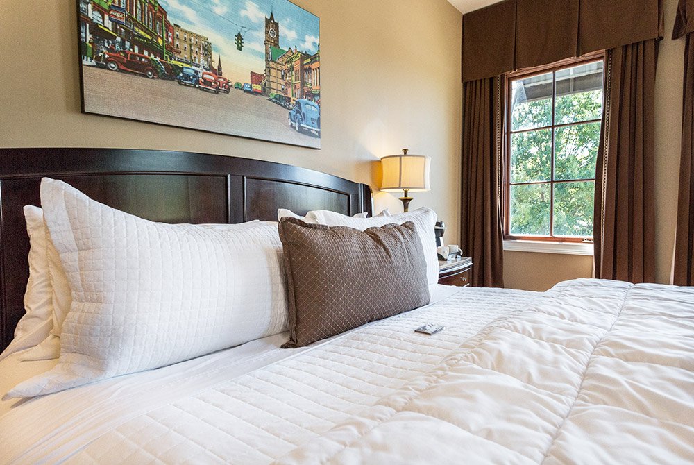 Hotel room with a bed featuring white quilted bedding and several pillows, including one with a brown geometric pattern, beneath a painting of a vintage street scene.