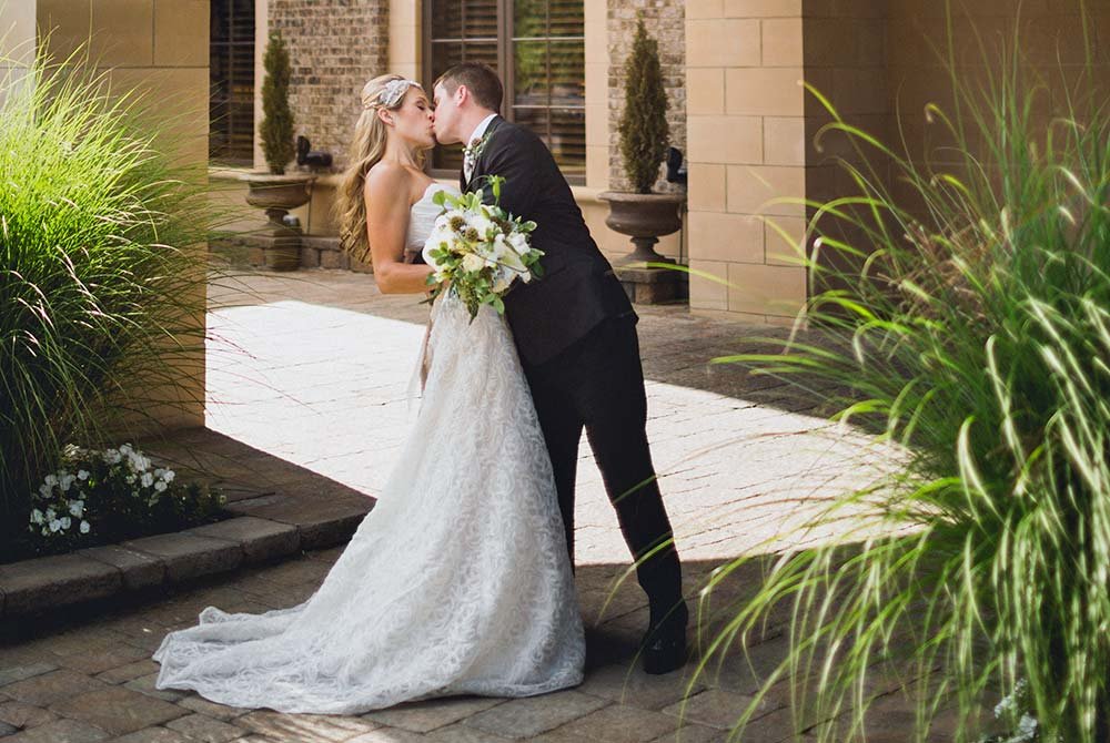 A bride in a white wedding gown and a groom in a dark suit kiss while standing on a stone patio.